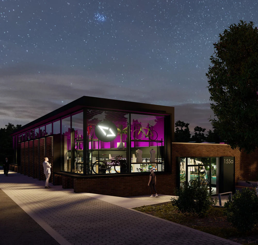 Fahrradgeschäft in modernem Gebäude bei Nacht, mit Glasfronten und pinker Innenbeleuchtung, Menschen davor.