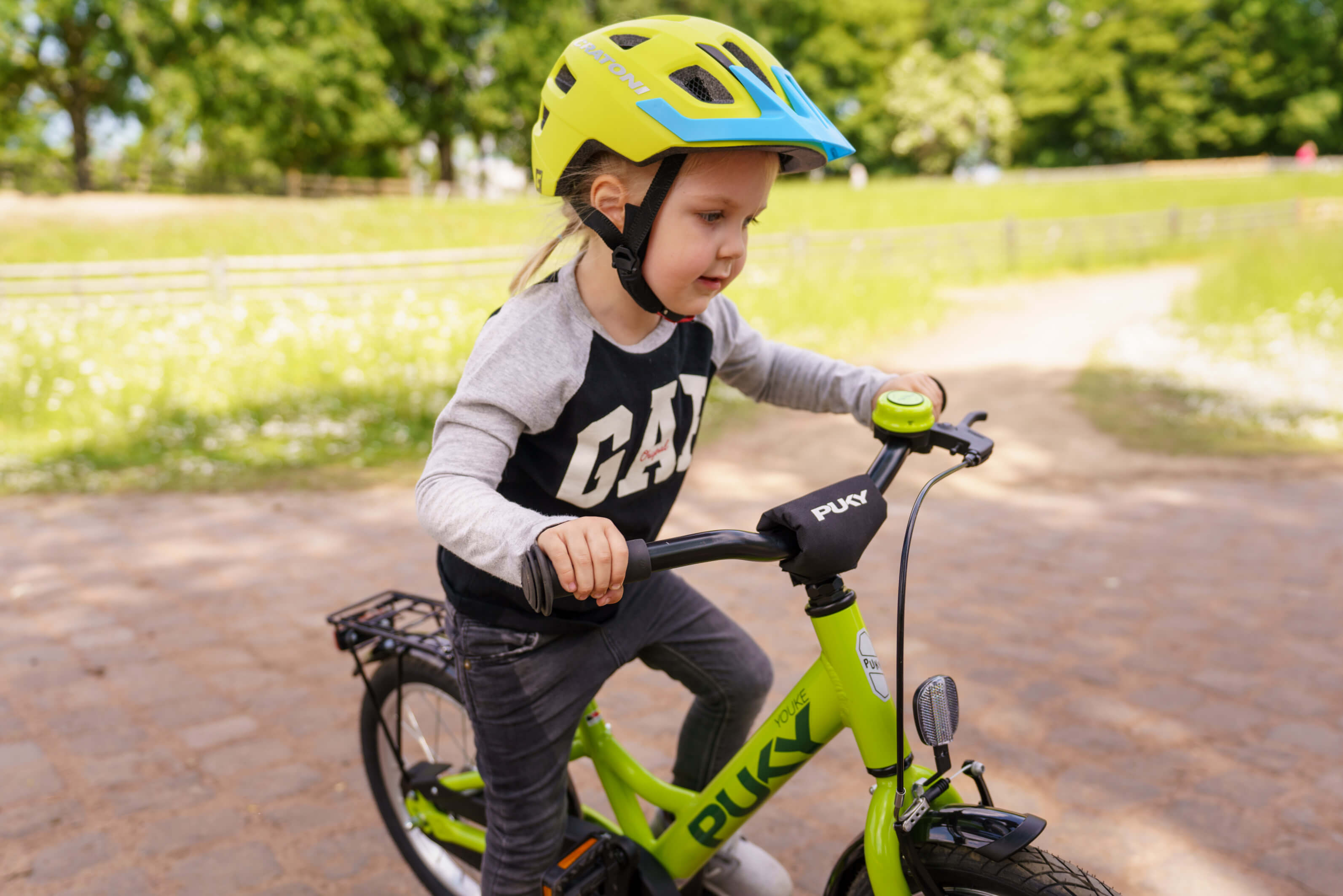 Kind auf grünem Puky Fahrrad, trägt gelb-blauen Helm. Geeignet für junge Radfahrer, im Freien.