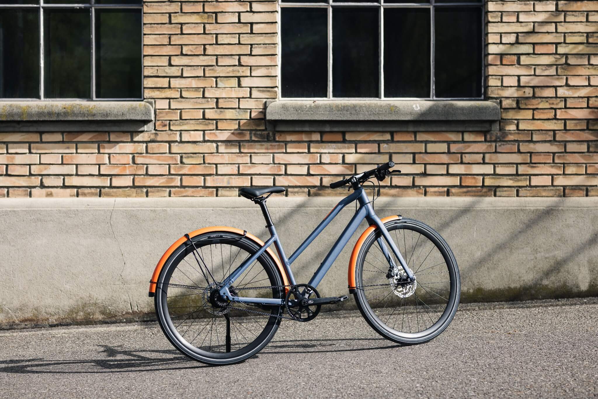 Graues BMC City-Bike mit orangenen Schutzblechen, lehnt an Ziegelwand. Ideal für urbane Pendler.