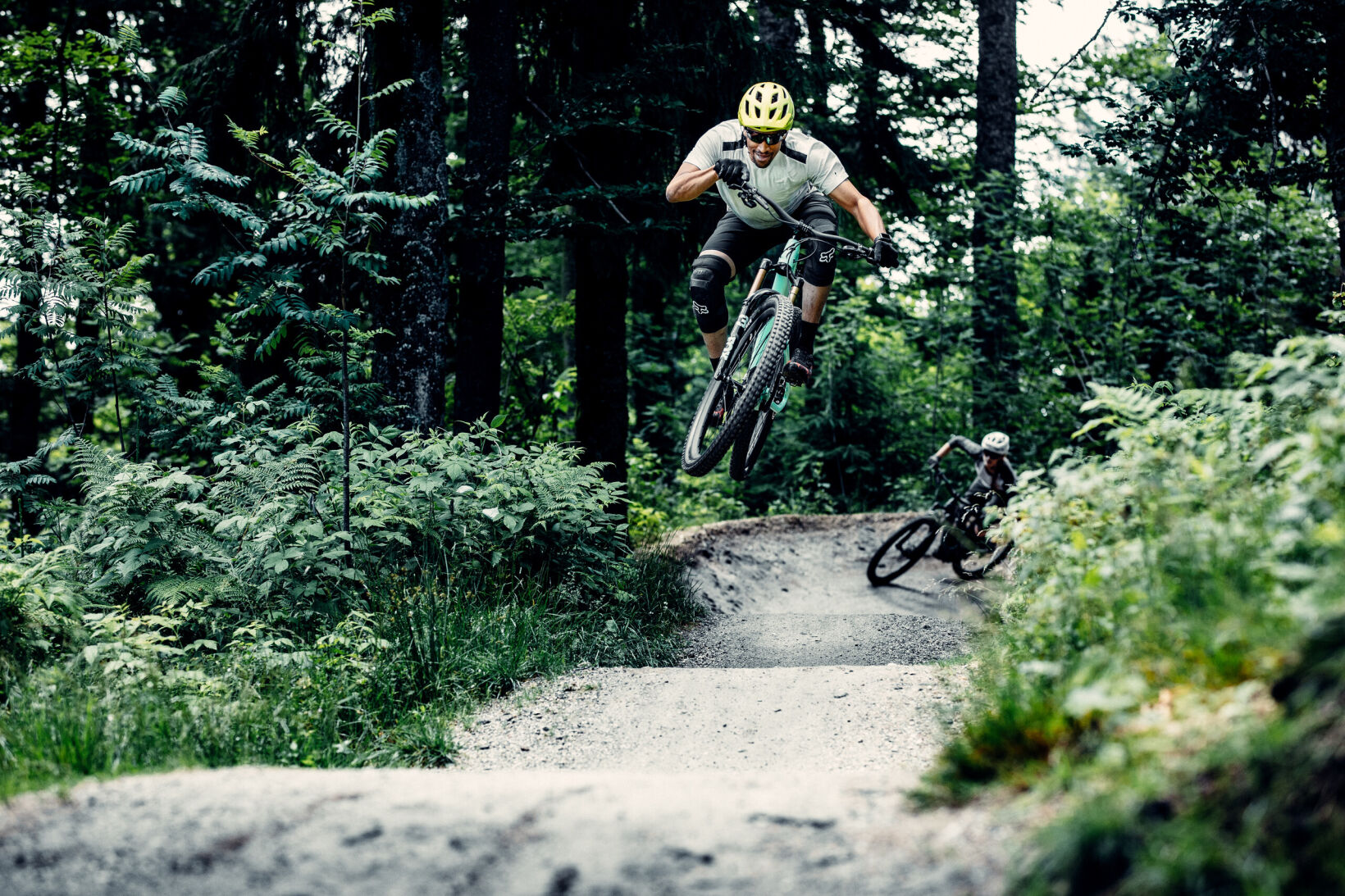 Zwei Mountainbiker springen auf einer Waldstrecke; einer in der Luft, beide mit Helmen und Schutzkleidung.