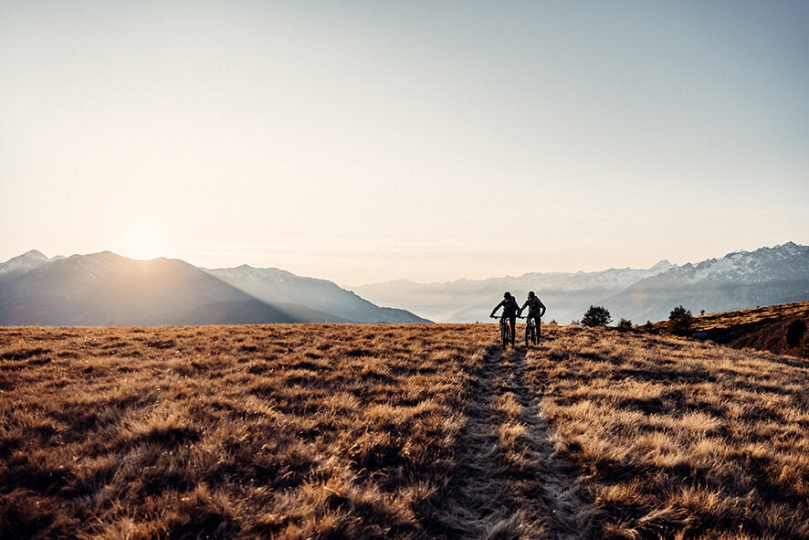 Zwei Mountainbiker fahren auf einem grasbewachsenen Pfad in einer weiten Berglandschaft bei Sonnenuntergang.
