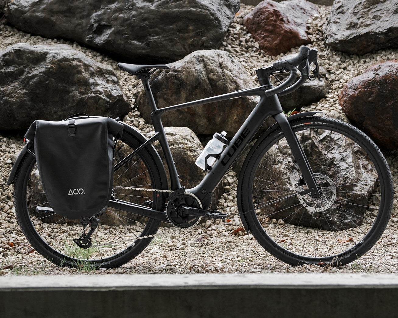 Schwarzes Cube Nuroad Hybrid Fahrrad mit Lenker, Rahmentasche und schwarzen Packtaschen vor steiniger Kulisse.