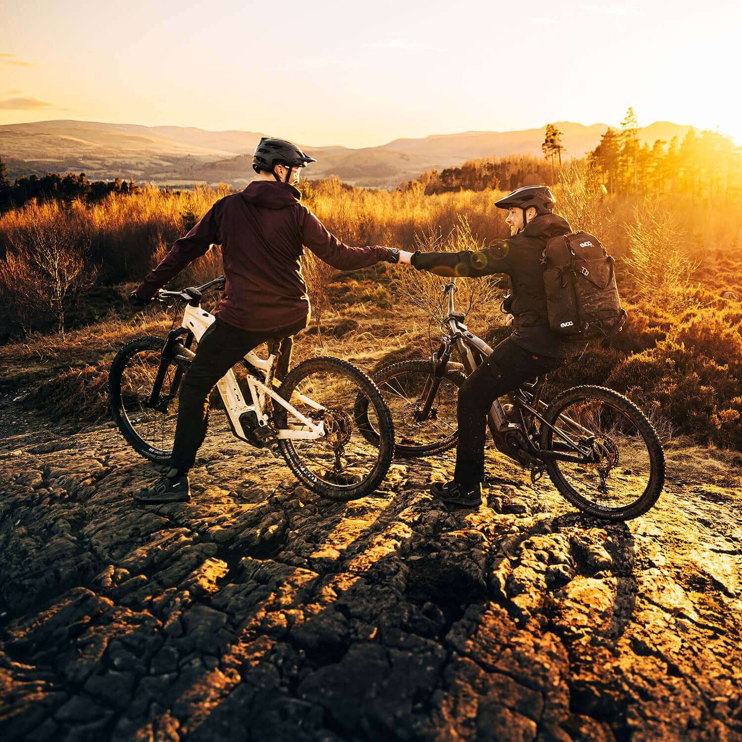 Zwei Mountainbiker mit Helmen geben sich Faustgruß bei Sonnenuntergang auf einem felsigen Weg.