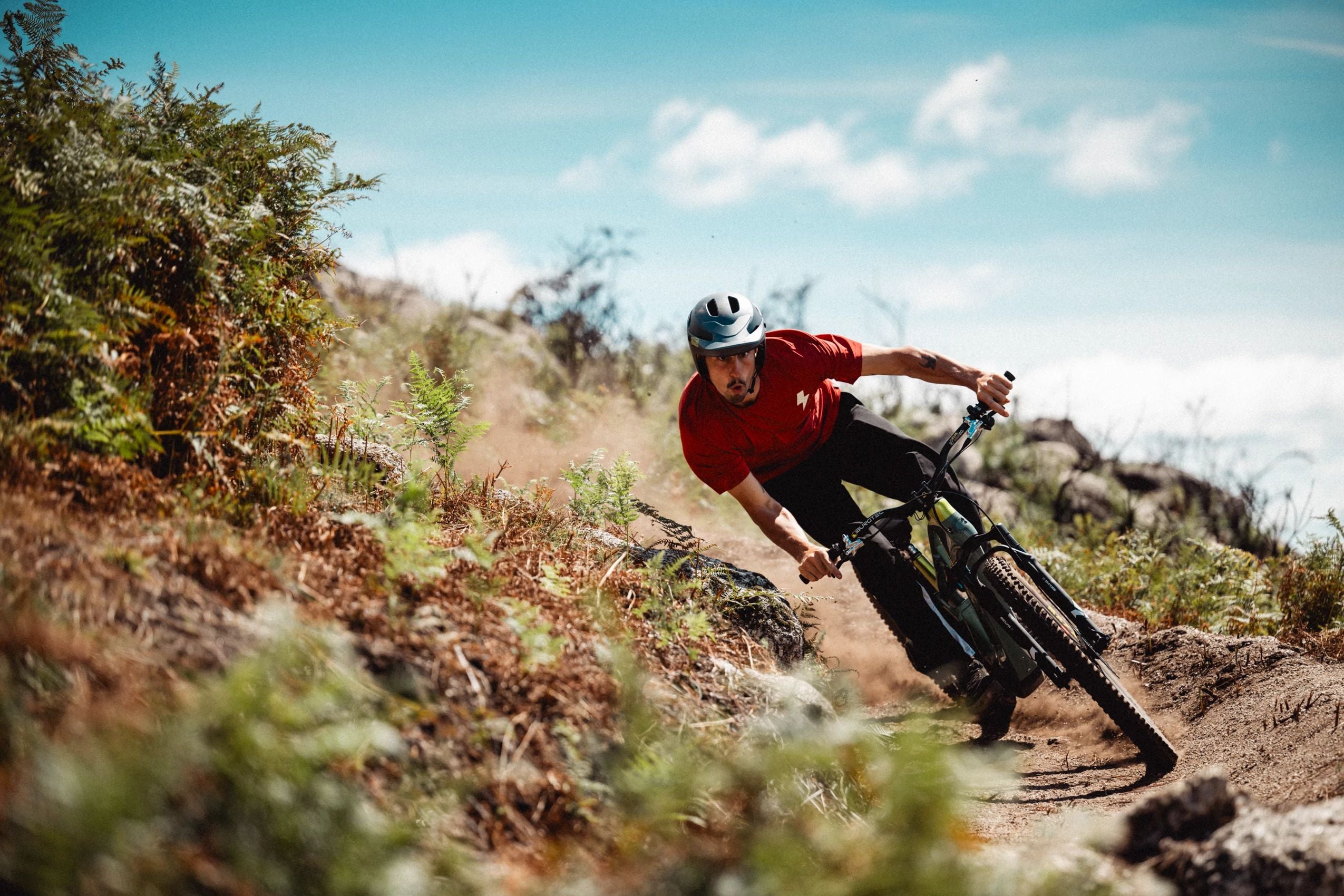 Mountainbiker auf Cannondale E-Bike, fährt schnell auf einem staubigen Trail, umgeben von Grün und blauem Himmel.
