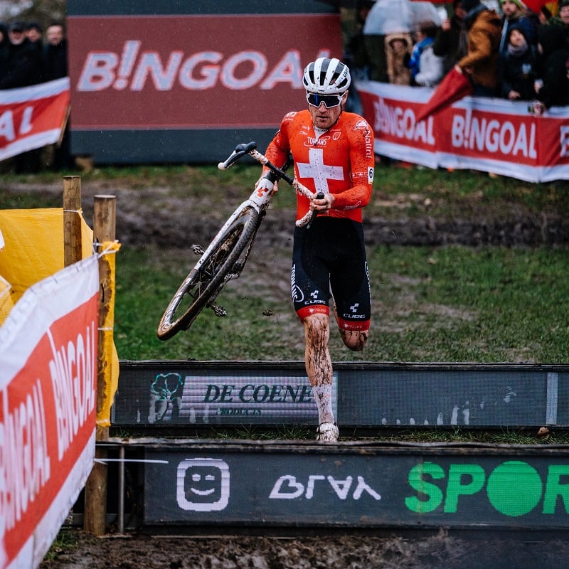 Radrennfahrer in rotem Trikot trägt sein Fahrrad bei einem cyclocross Rennen durch matschigen Parcours.