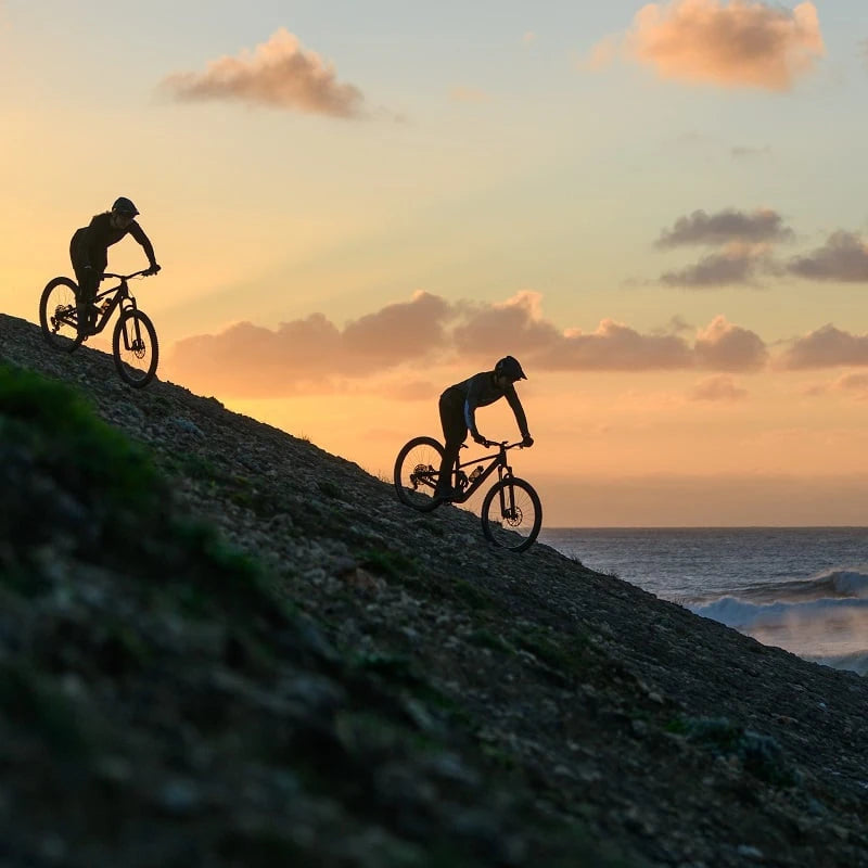 Zwei Mountainbiker fahren bei Sonnenuntergang einen Hügel hinunter.