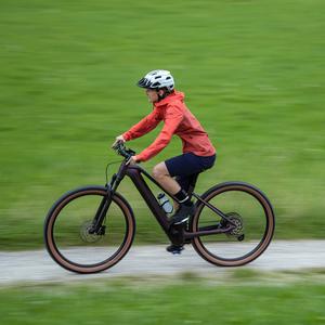 Person fährt ein Cube E-Gravelbike auf Kiesweg, rote Jacke, Helm, grüne Wiese im Hintergrund. Ideal für sportliche Radfahrer*innen.
