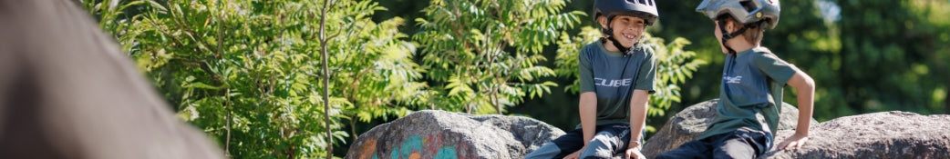 Kinder tragen Cube T-Shirts und Helme, sitzen auf Steinen in der Natur. Geeignet für Outdoor-Aktivitäten und Radsport.