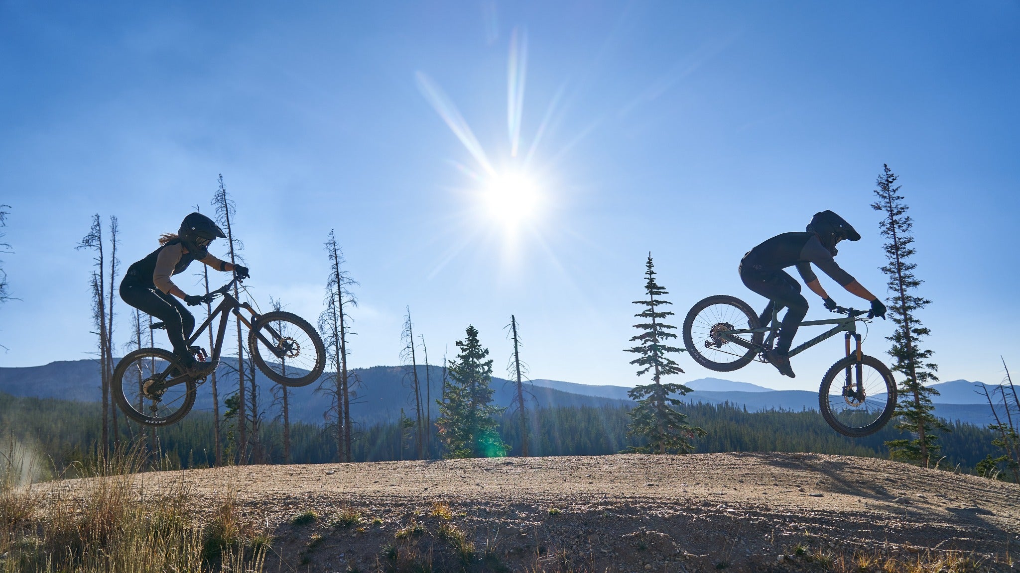 Zwei Mountainbiker springen mit Pivot Mountainbikes in bergiger Landschaft, blauer Himmel, helle Sonne im Hintergrund.