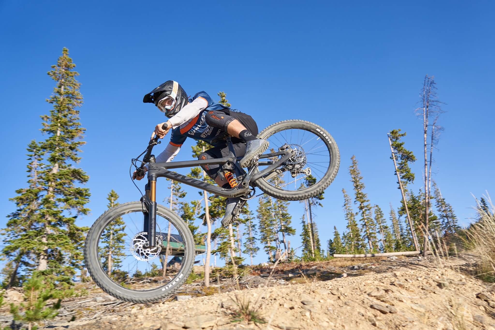 Mountainbiker auf Pivot Mach 6 Bike springt auf Waldpfad vor blauem Himmel. Ideal für Trail-Enthusiasten.