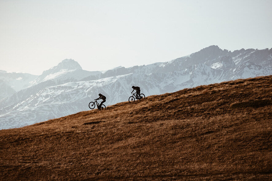 Zwei Radfahrer mit E-Mountainbikes fahren eine Wiese im Gebirge hinauf, Berge im Hintergrund. Ideal für Outdoor-Abenteuer.