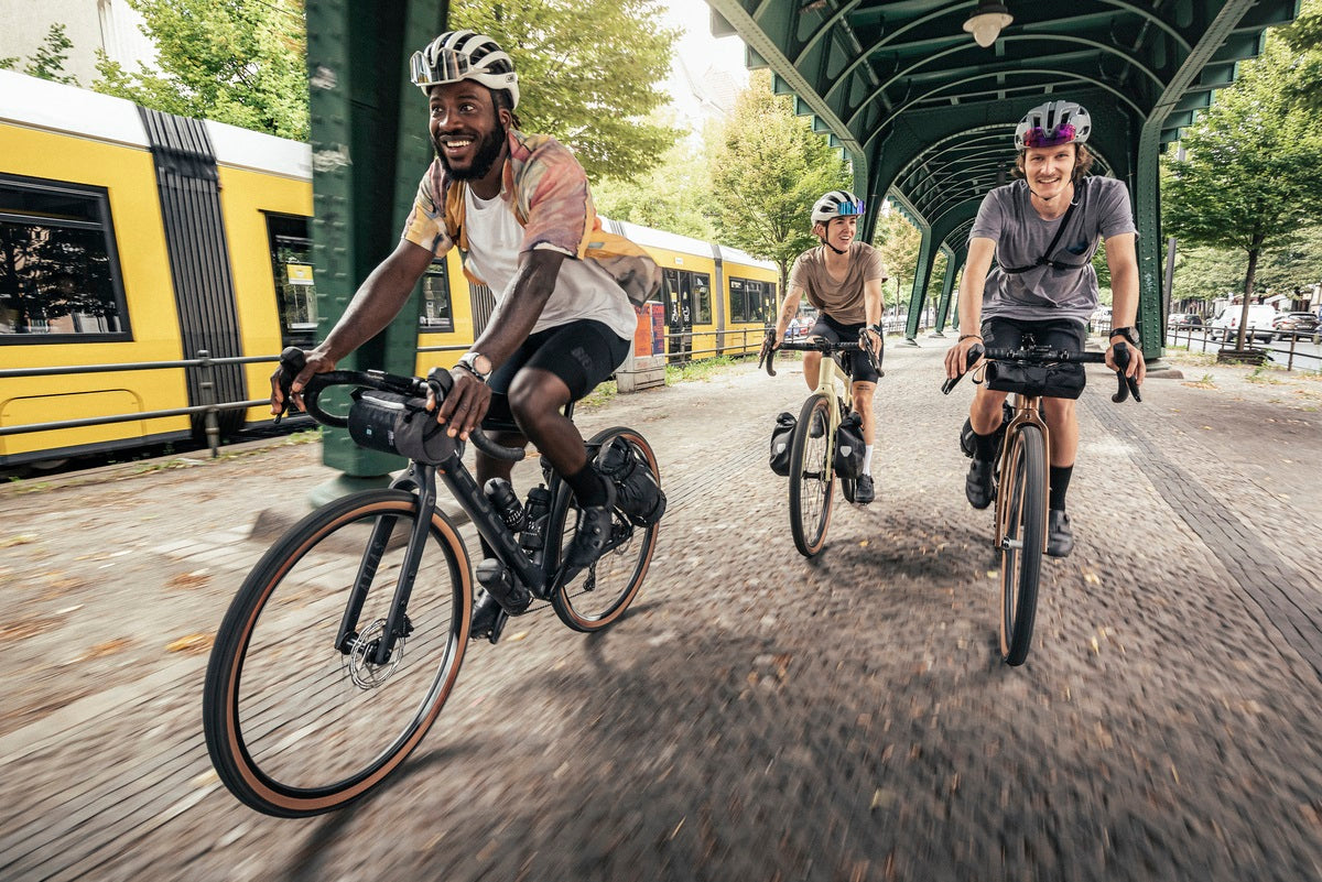 Drei Personen fahren unter einer Brücke auf Gravel-Bikes der Marke Focus, Modell Atlas; Straßenbahn im Hintergrund.