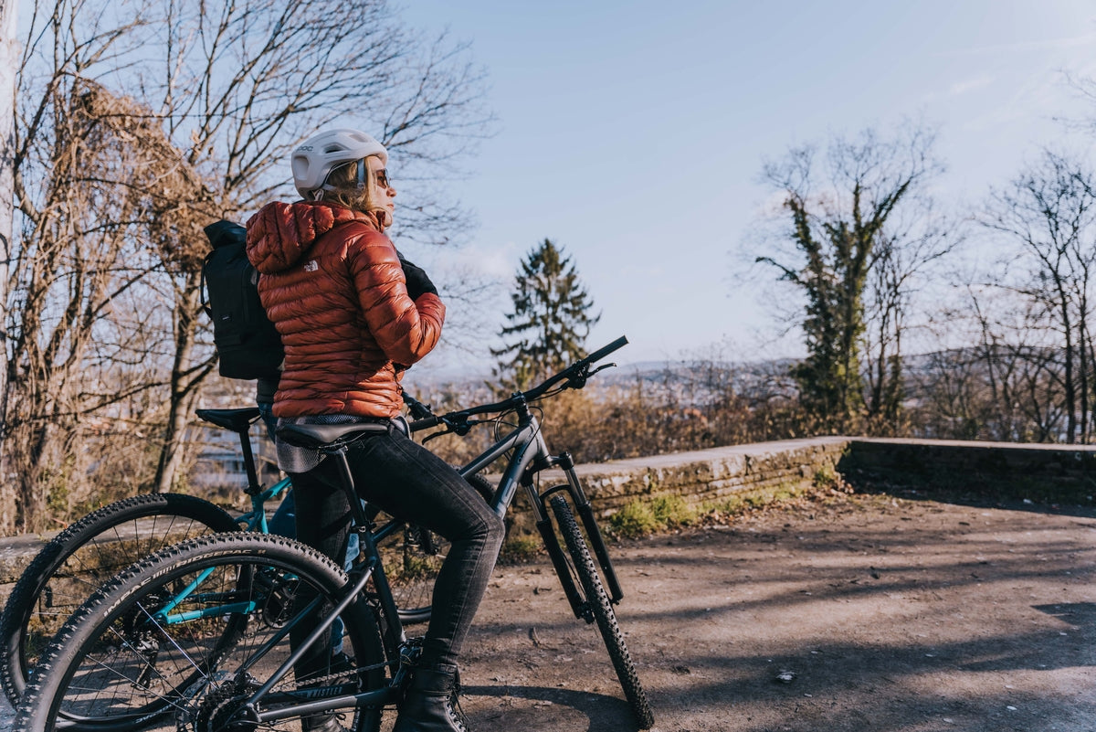 Person auf einem Mountainbike im Freien mit Herbstlandschaft, rote Jacke, Helm, zwei Fahrräder sichtbar.
