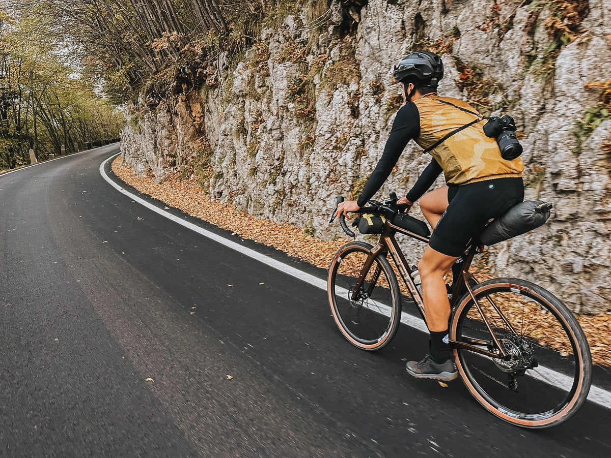 Fahrradfahrer auf einem Gravelbike fährt mit Kamera-Ausrüstung auf einer kurvigen bergigen Straße in herbstlicher Landschaft.