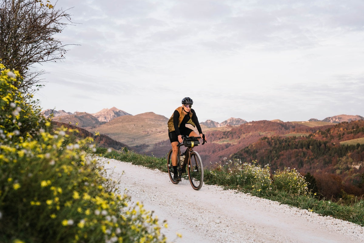Radfahrer mit Helm auf einem Gravelbike auf einem Schotterweg in hügeliger Landschaft bei Tageslicht.
