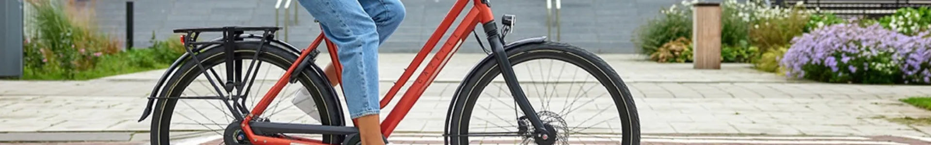 Person fährt rotes Gazelle-Fahrrad, seitliche Ansicht. Trägt Jeans und Sneaker. Stadtumgebung im Hintergrund.