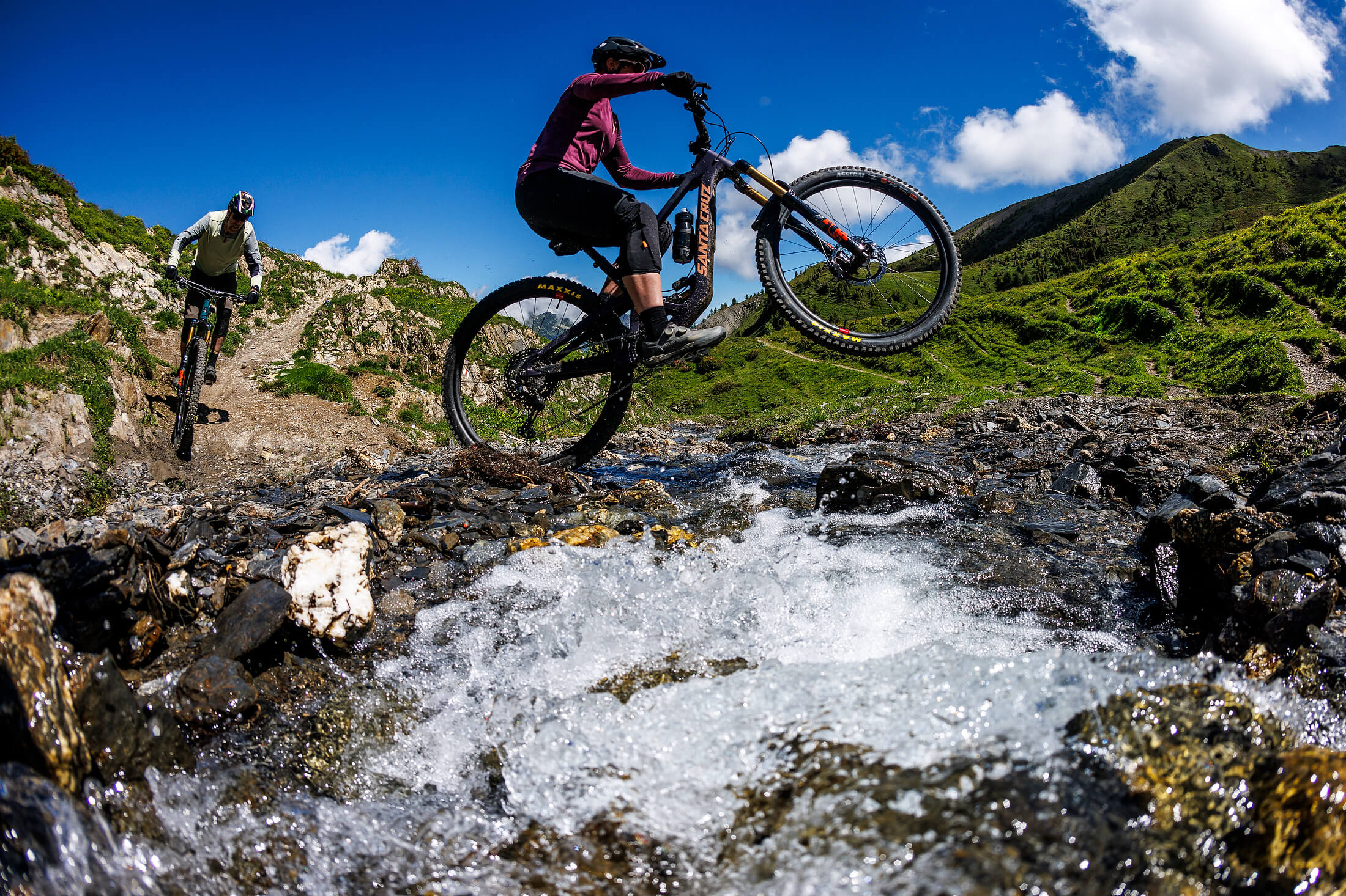 Zwei Radfahrer auf Santa Cruz Hightower Mountainbikes überqueren einen Gebirgsbach auf einer steinigen Strecke.