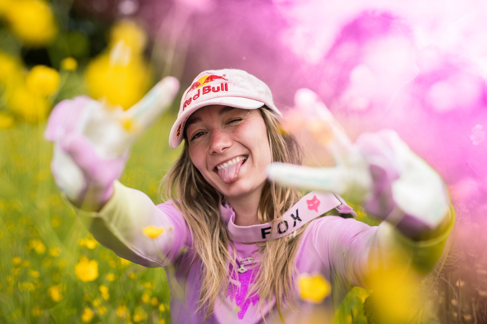 Junge Frau mit Red Bull Kappe, pinkem Oberteil und FOX-Gurt in einer blumigen Landschaft, zeigt ein verspieltes Gesicht.