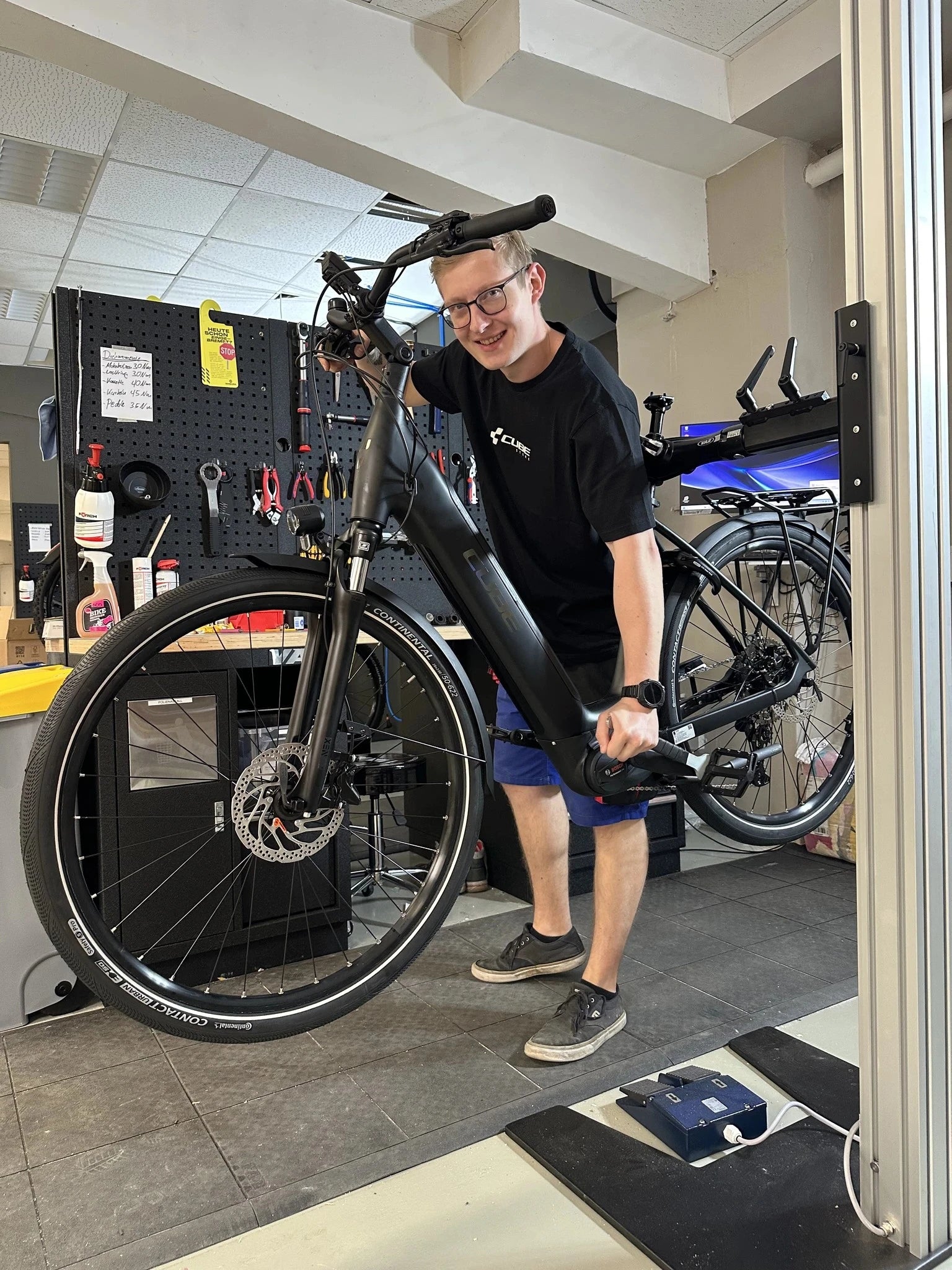 Mechaniker hebt ein schwarzes Cube E-Bike in Werkstatt hoch, umgeben von Werkzeugen und Fahrradzubehör.