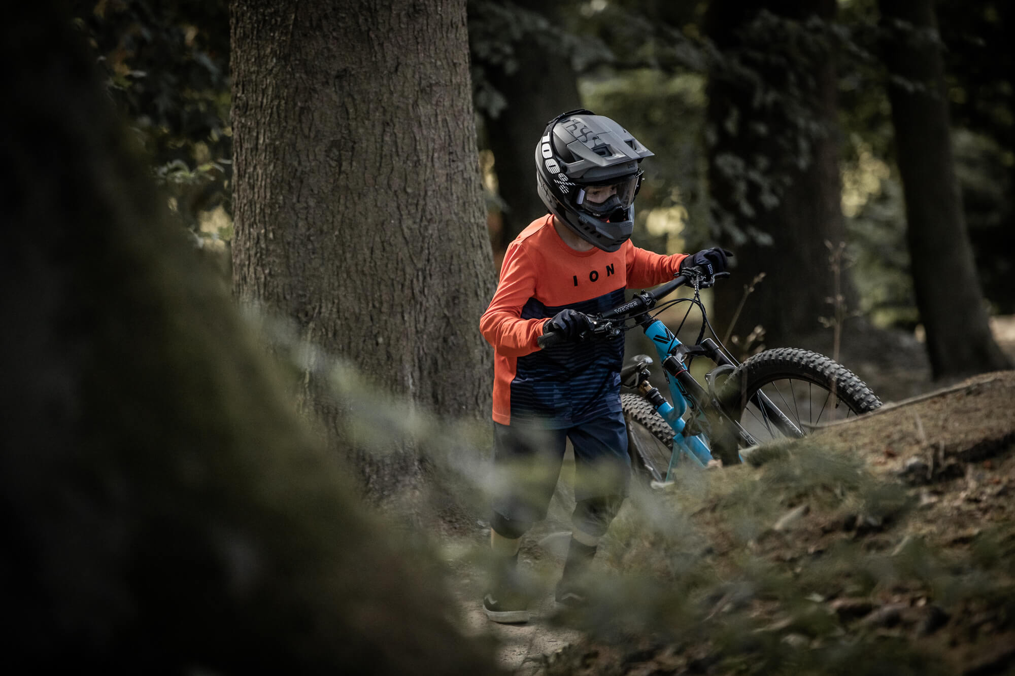 Kind in ION Fahrradtrikot und Helm schiebt Mountainbike im Wald.