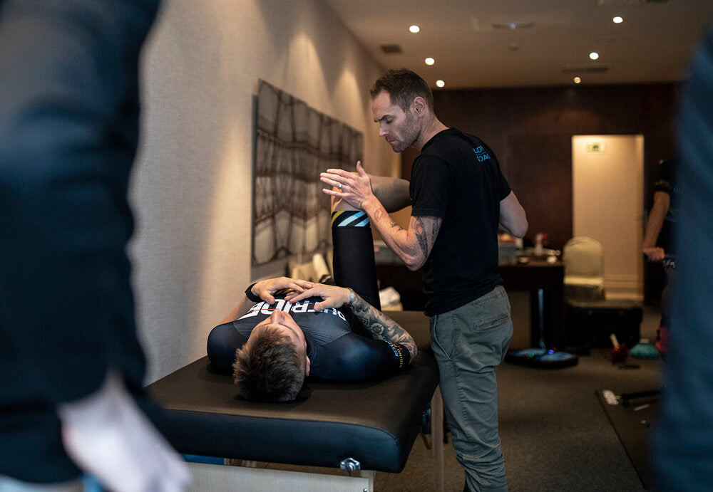 Ein Therapeut führt in einem Raum eine Dehnübung an einem Athleten auf einer Liege durch.