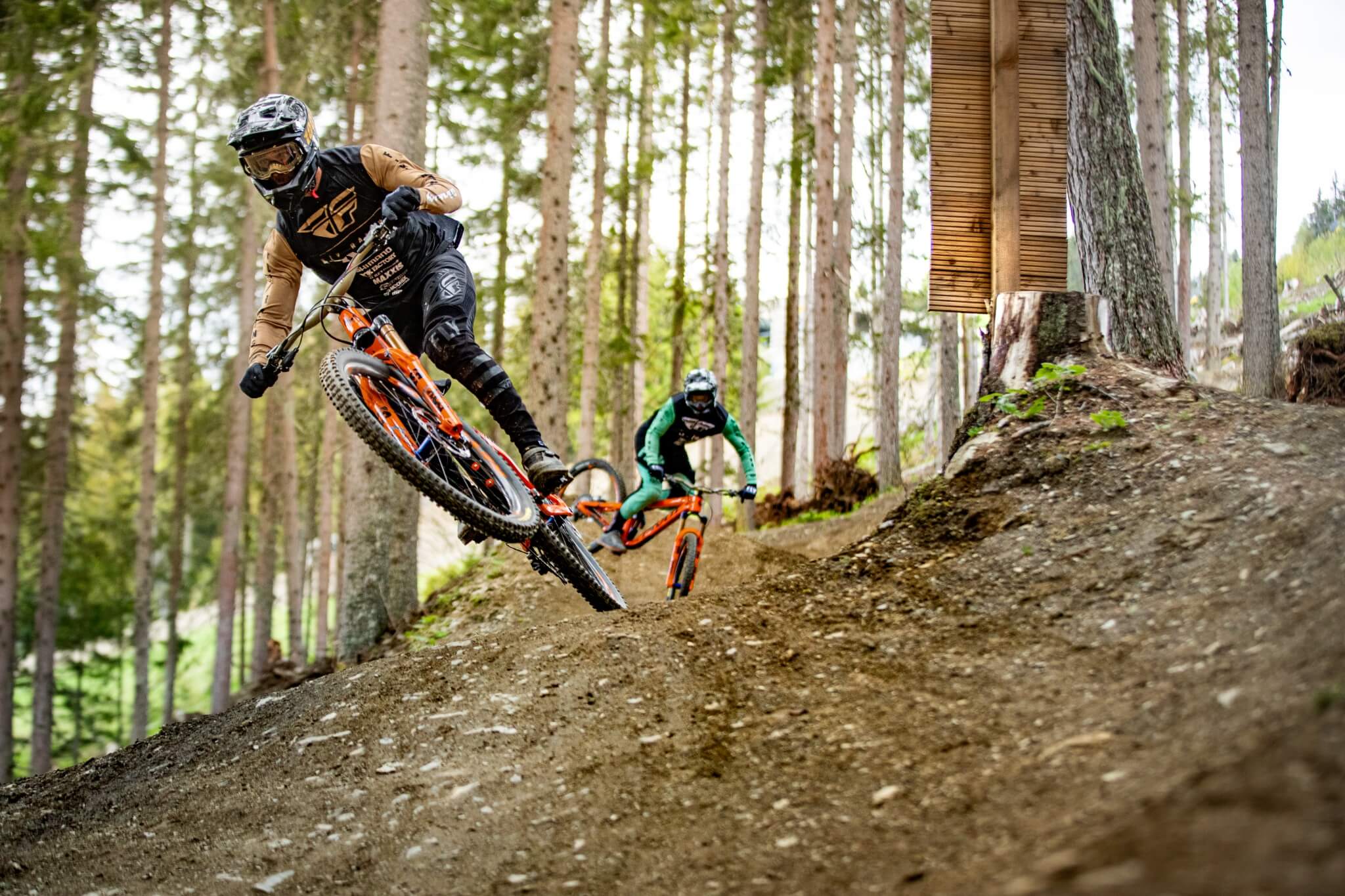 Zwei Mountainbiker fahren auf orangefarbenen Fahrrädern durch einen Wald mit Helm und Schutzkleidung.