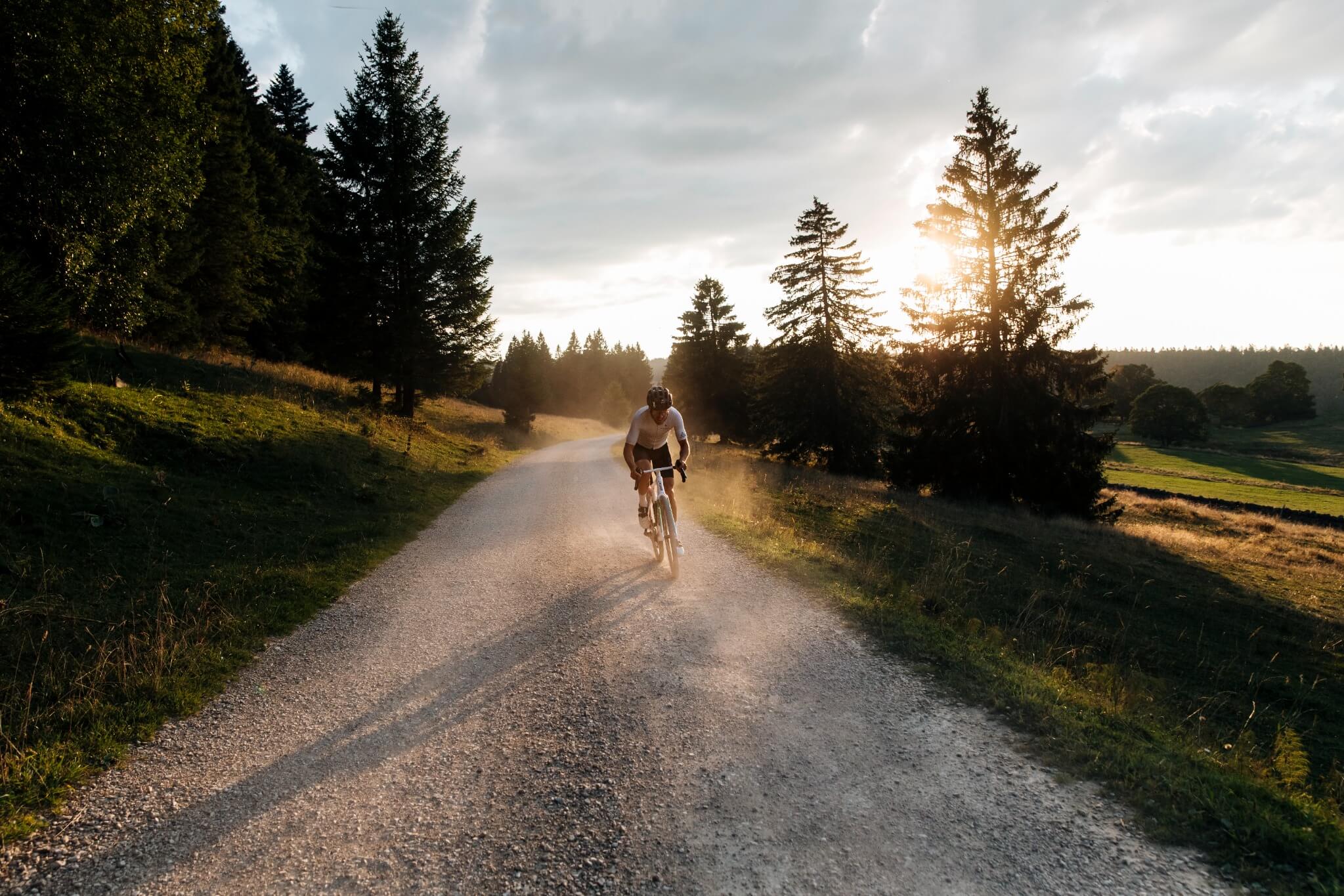 Radfahrer fährt bei Sonnenuntergang auf einer Schotterstraße, umgeben von Waldlandschaft.