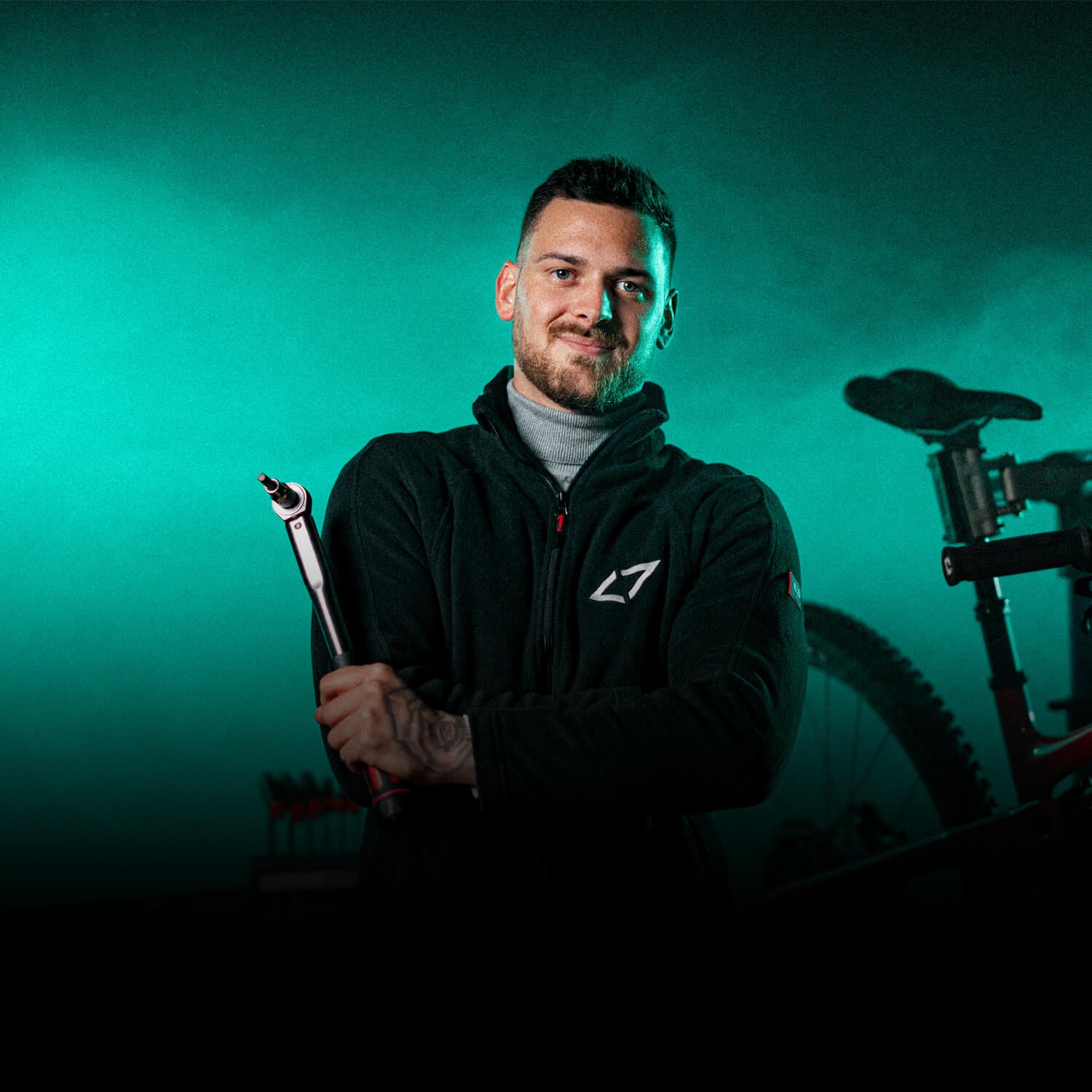 Mann in schwarzer Jacke mit Logo hält Fahrradpumpe, im Hintergrund Mountainbike und grün-blauer Nebel.