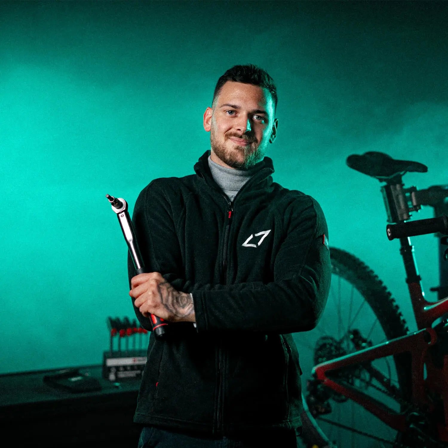 Ein Mann hält ein Werkzeug vor einem Fahrrad, im Hintergrund Werkbank mit Werkzeugen, trägt L7-Jacke.