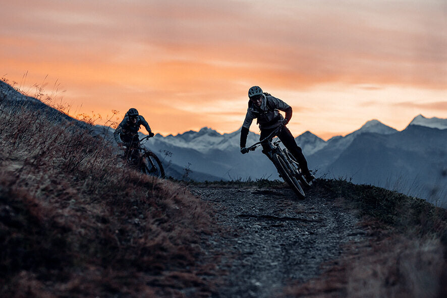 Zwei Mountainbiker fahren bei Sonnenuntergang einen Bergpfad hinunter.