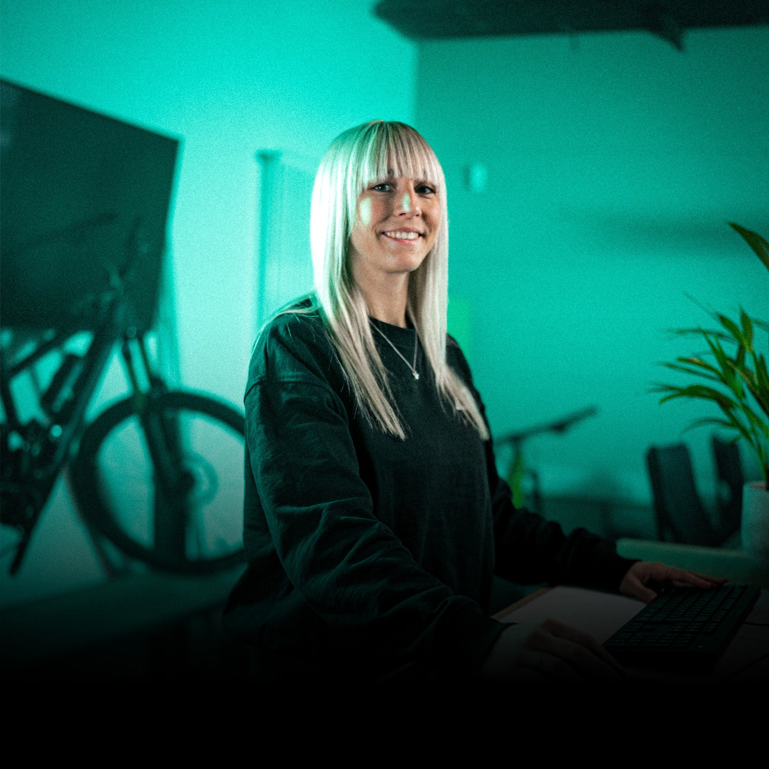 Eine Frau arbeitet an einem Schreibtisch mit Computer, Hintergrund unscharf mit Fahrrad und Pflanze.