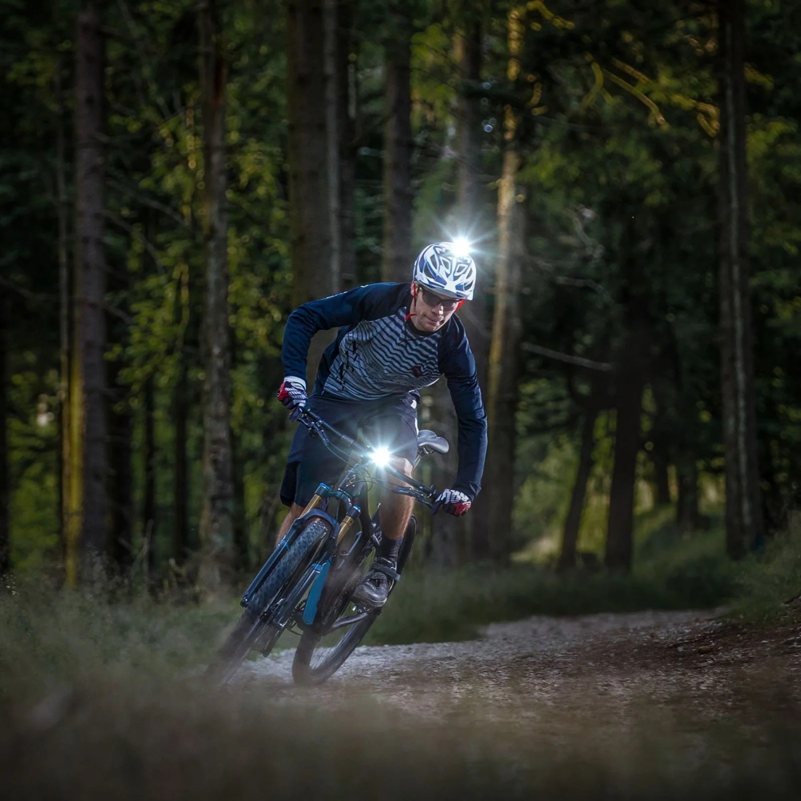 Radfahrer mit Helm- und Frontlicht auf Waldweg bei Dämmerung, geeignet für Nachtfahrten; Marke und Modell nicht erkenntlich.