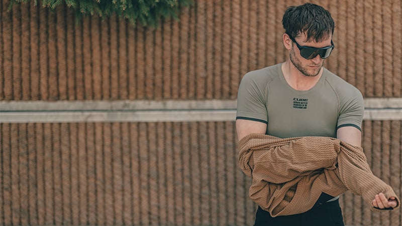 Mann im Cube Sportshirt, hält Jacke, trägt Sonnenbrille, steht vor Seilwand.