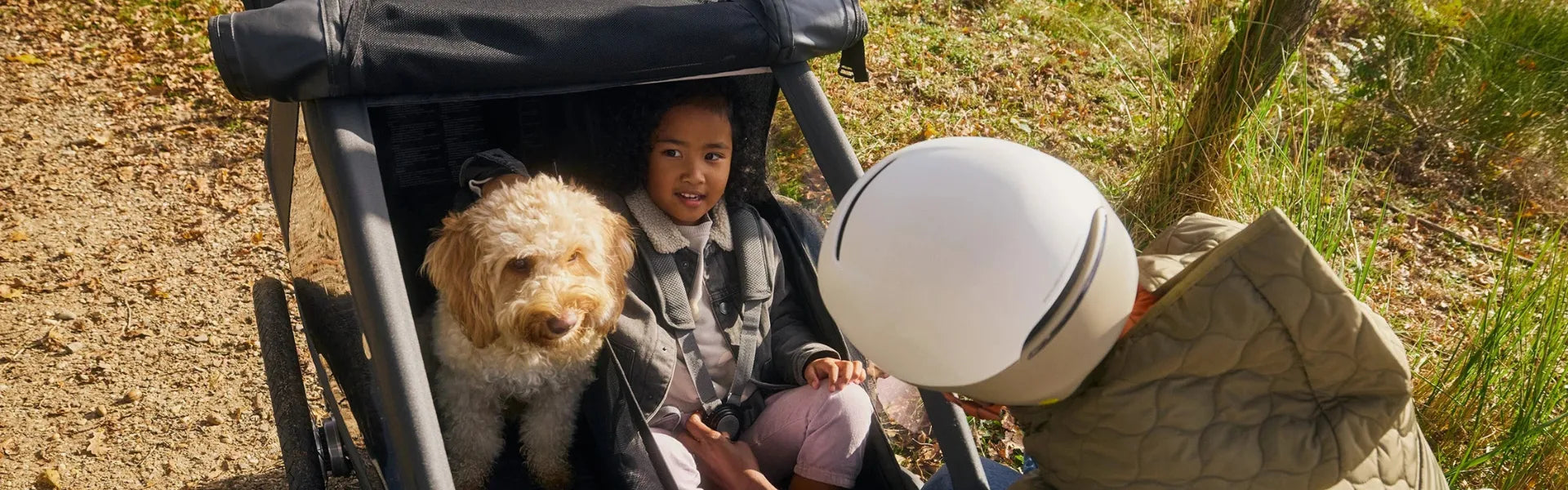 Kind und Hund in einem Croozer Fahrradanhänger für Kinder auf einem Waldweg, daneben ein Erwachsener mit Helm. Ideal für Familienausflüge.
