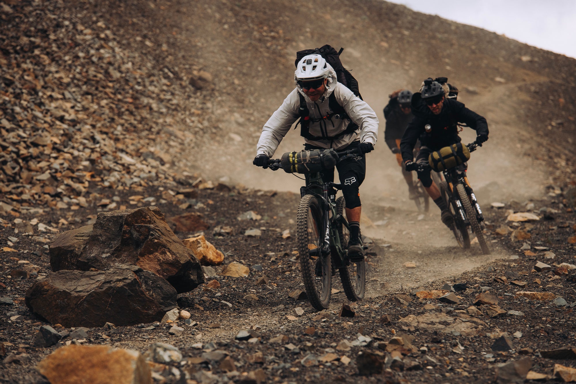 Drei Mountainbiker fahren in Schutzkleidung auf einem steinigen Weg bergab, geeignet für Outdoor-Abenteuer.