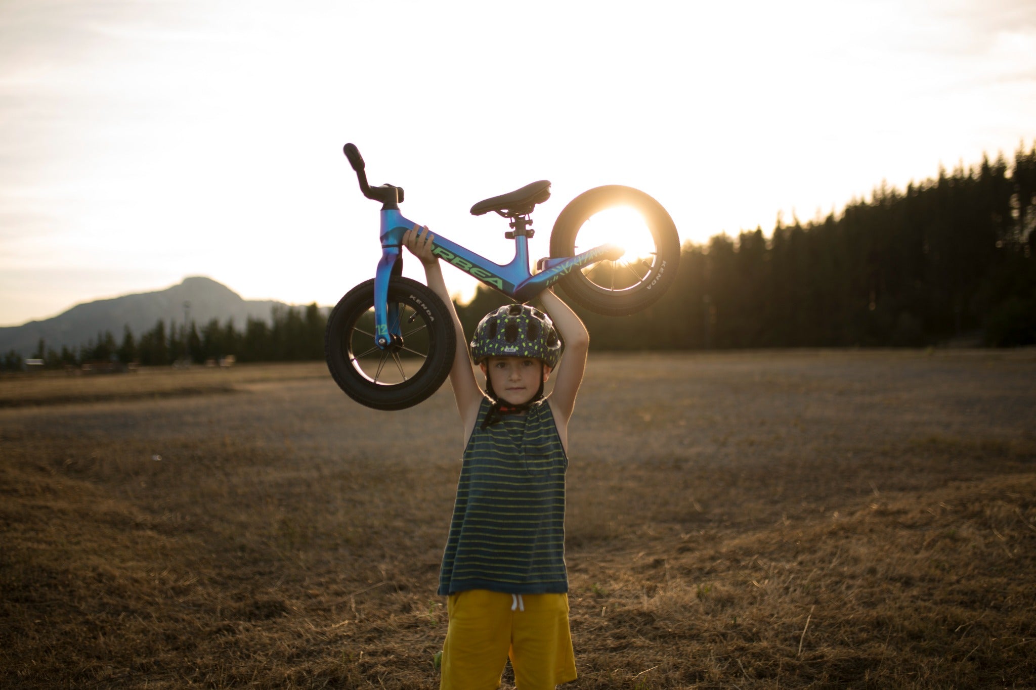 Kind hebt ein blaues Orbea-Laufrad über Kopf, trägt Helm, natürliche Landschaft im Sonnenuntergang. Ideal für junge Anfänger.