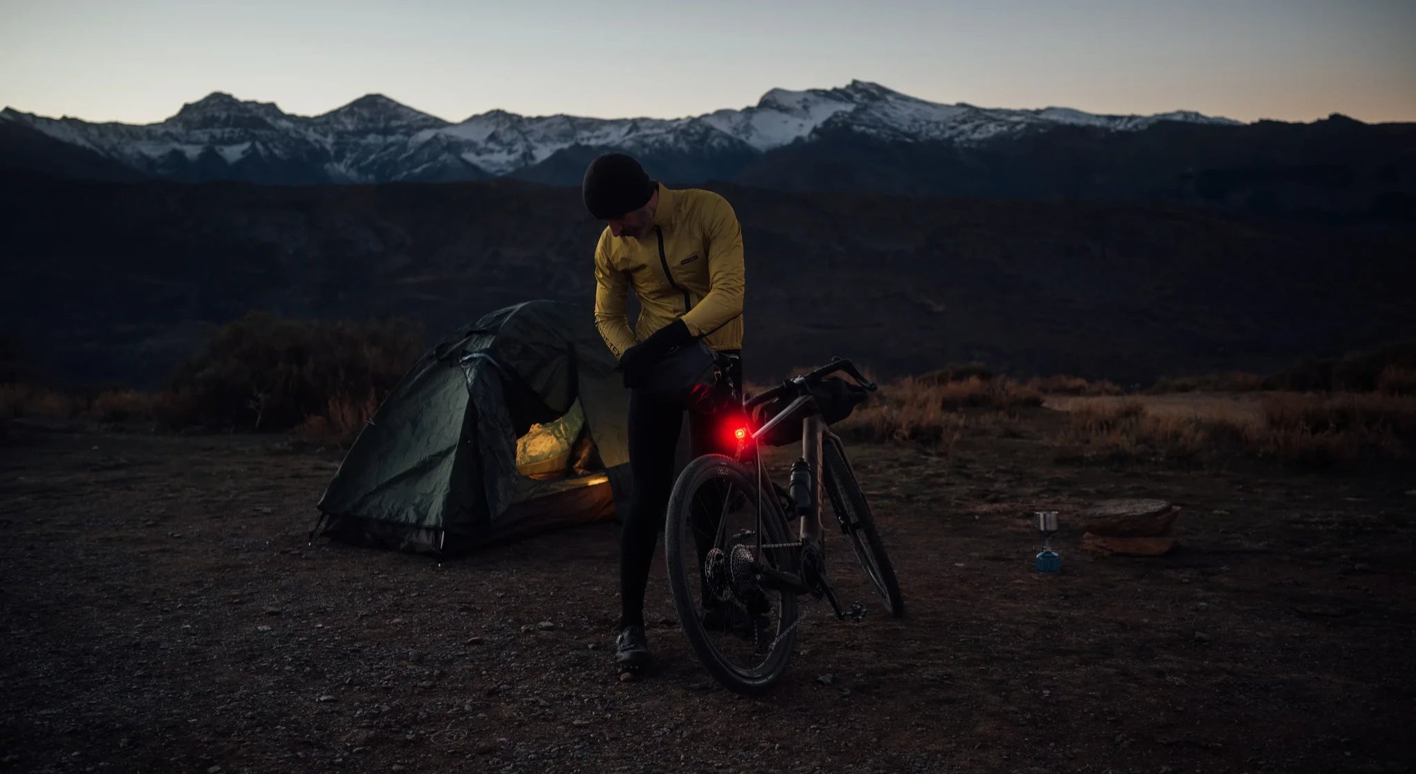 Mann in gelber Jacke neben Zelt und Fahrrad bei Dämmerung in den Bergen, vorbereitet für Campingausflug.