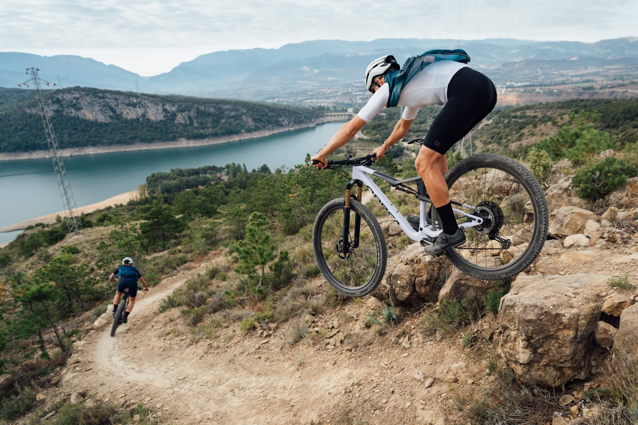 Zwei Mountainbiker auf einem Trail mit Orbea Oiz Bikes, in bergiger Landschaft mit See im Hintergrund.