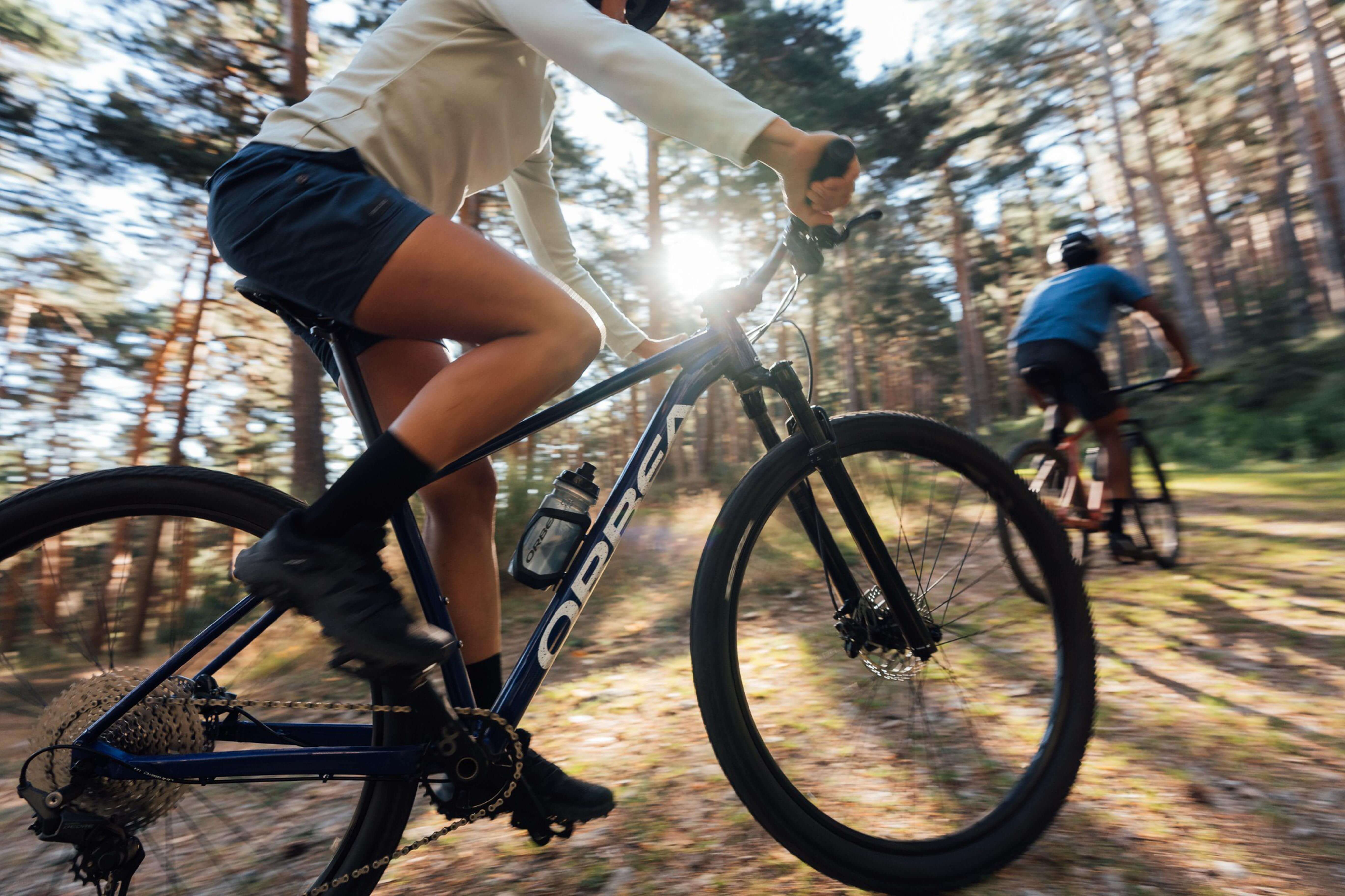 Zwei Radfahrer fahren mit Orbea Hardtail-Mountainbikes auf einem Waldweg im Sonnenschein.