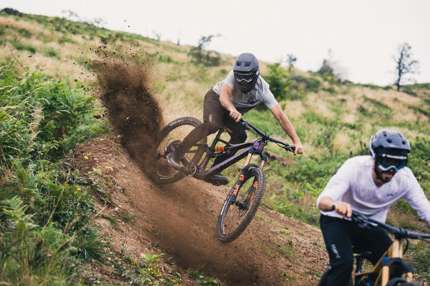 Zwei Mountainbiker in Aktion auf einem Trail, beide tragen Schutzausrüstung. Fahrrad: Orbea Rallon.