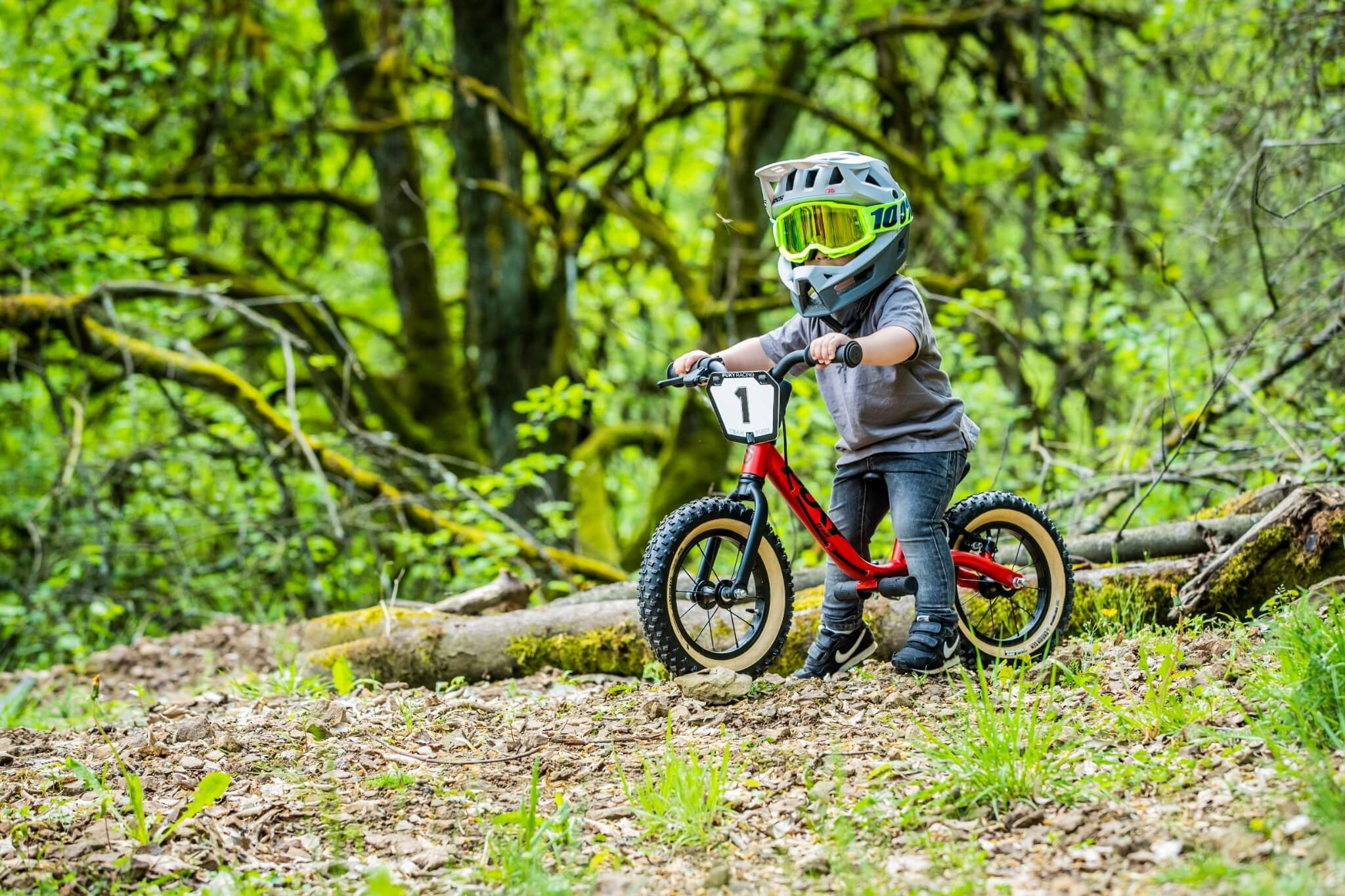Kind auf rotem Laufrad im Wald, trägt Helm und Schutzbrille. Ideal für Outdoor-Abenteuer.