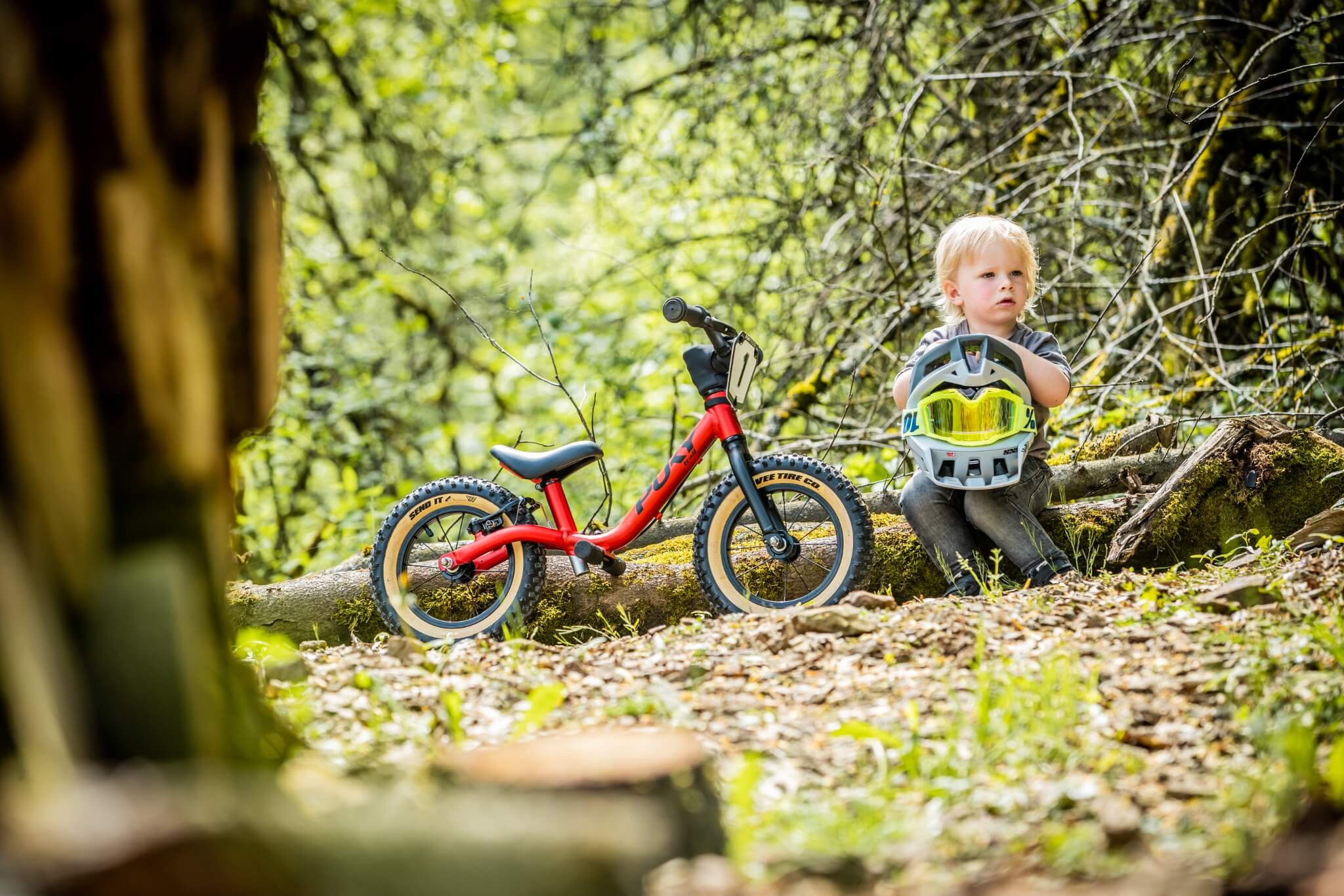 Rotes Kinderlaufrad von Yedoo im Wald, daneben ein Kind mit Helm sitzend. Ideal für junge Abenteurer draußen.