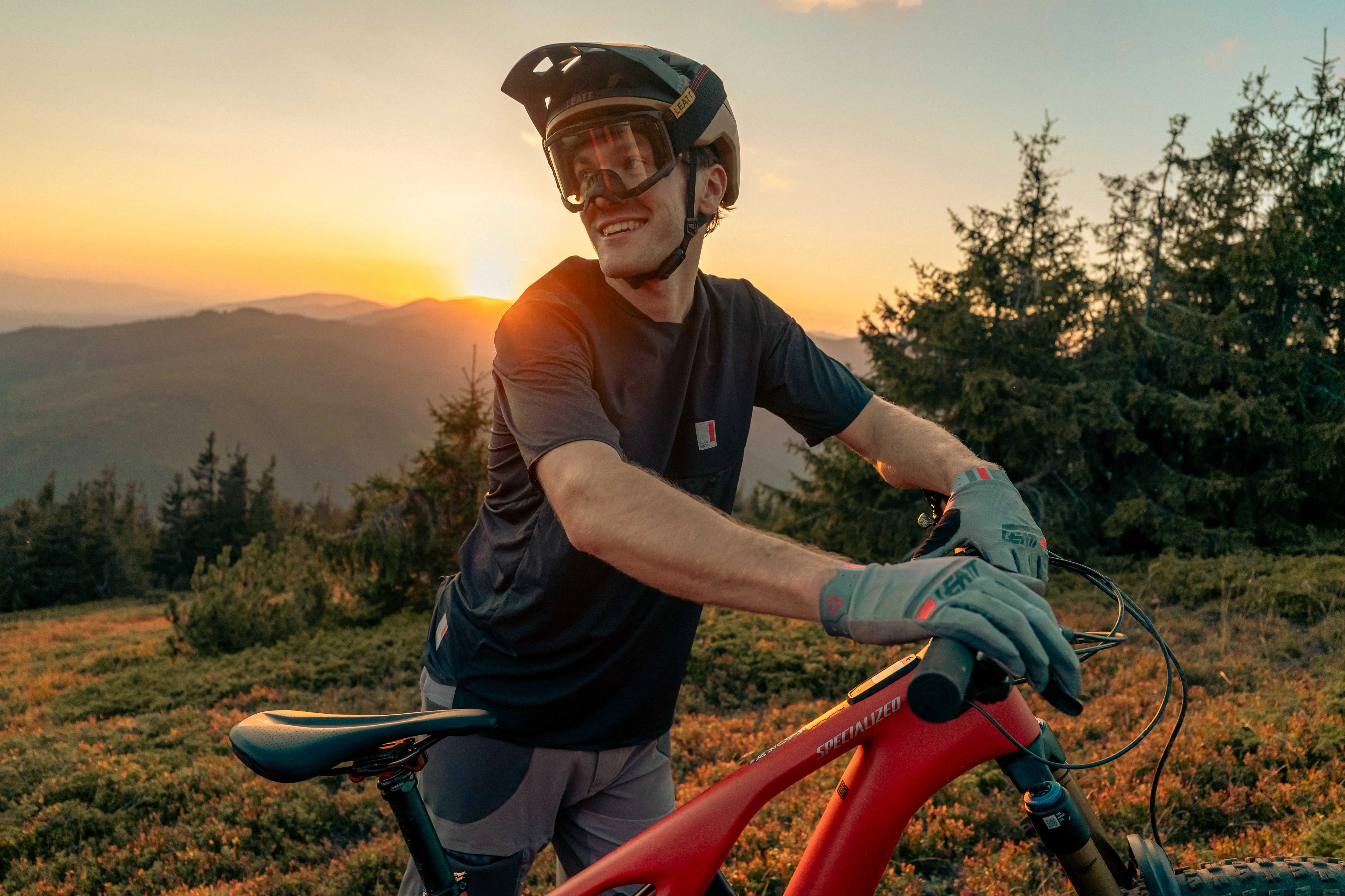 Mann mit Specialized-Mountainbike bei Sonnenuntergang, trägt Helm und Handschuhe, ideal für Mountainbike-Enthusiasten.