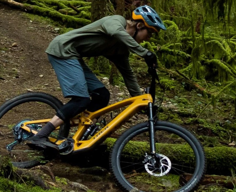 Mountainbiker auf gelbem Trek E-Bike im Wald, trägt Helm und Schutzkleidung, fährt auf einem schmalen Pfad.