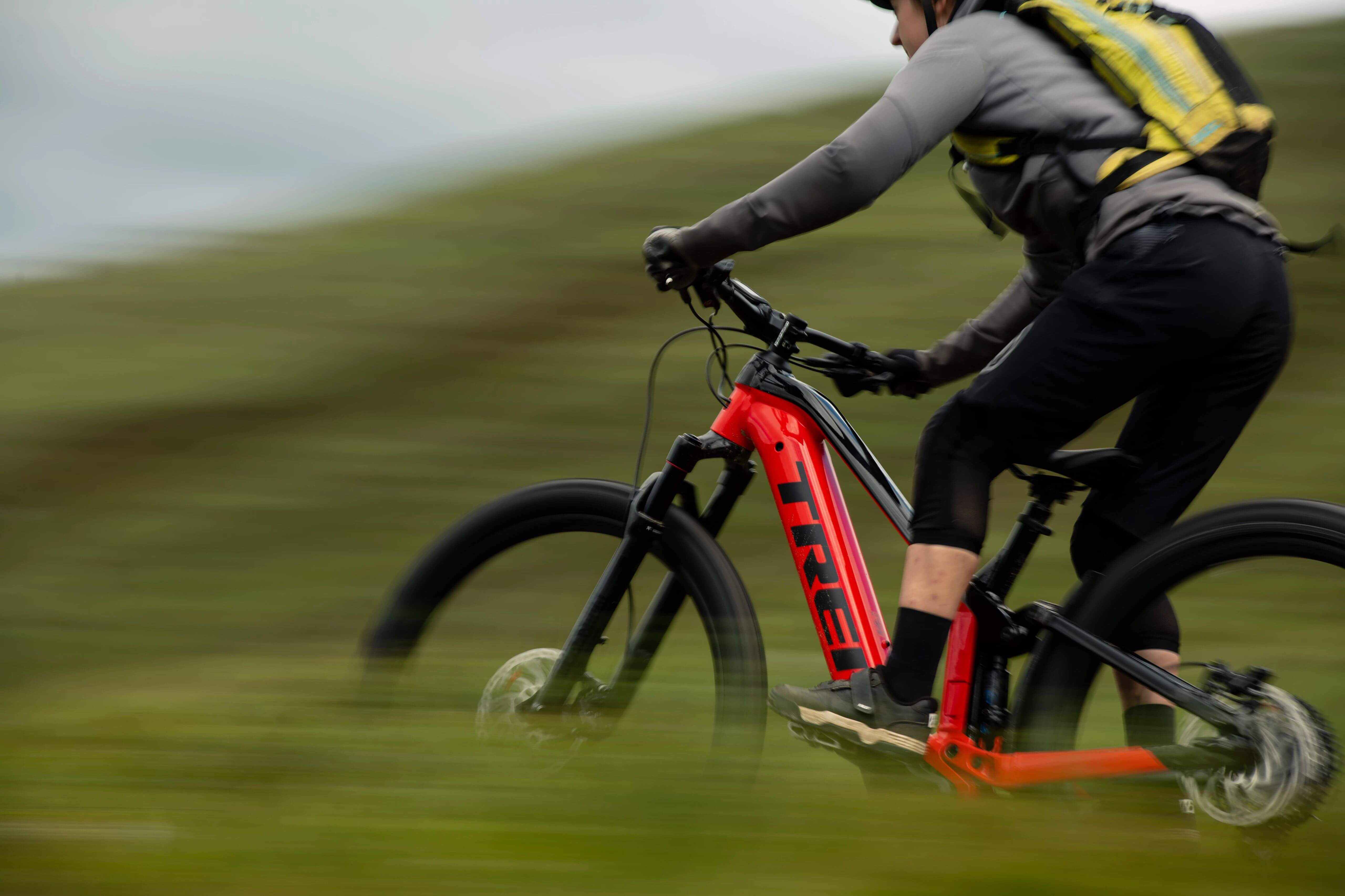 Rotes Trek E-Mountainbike Fullys fährt durch grüne Landschaft, Person mit Rucksack und Helm. Ideal für Offroad-Abenteuer.
