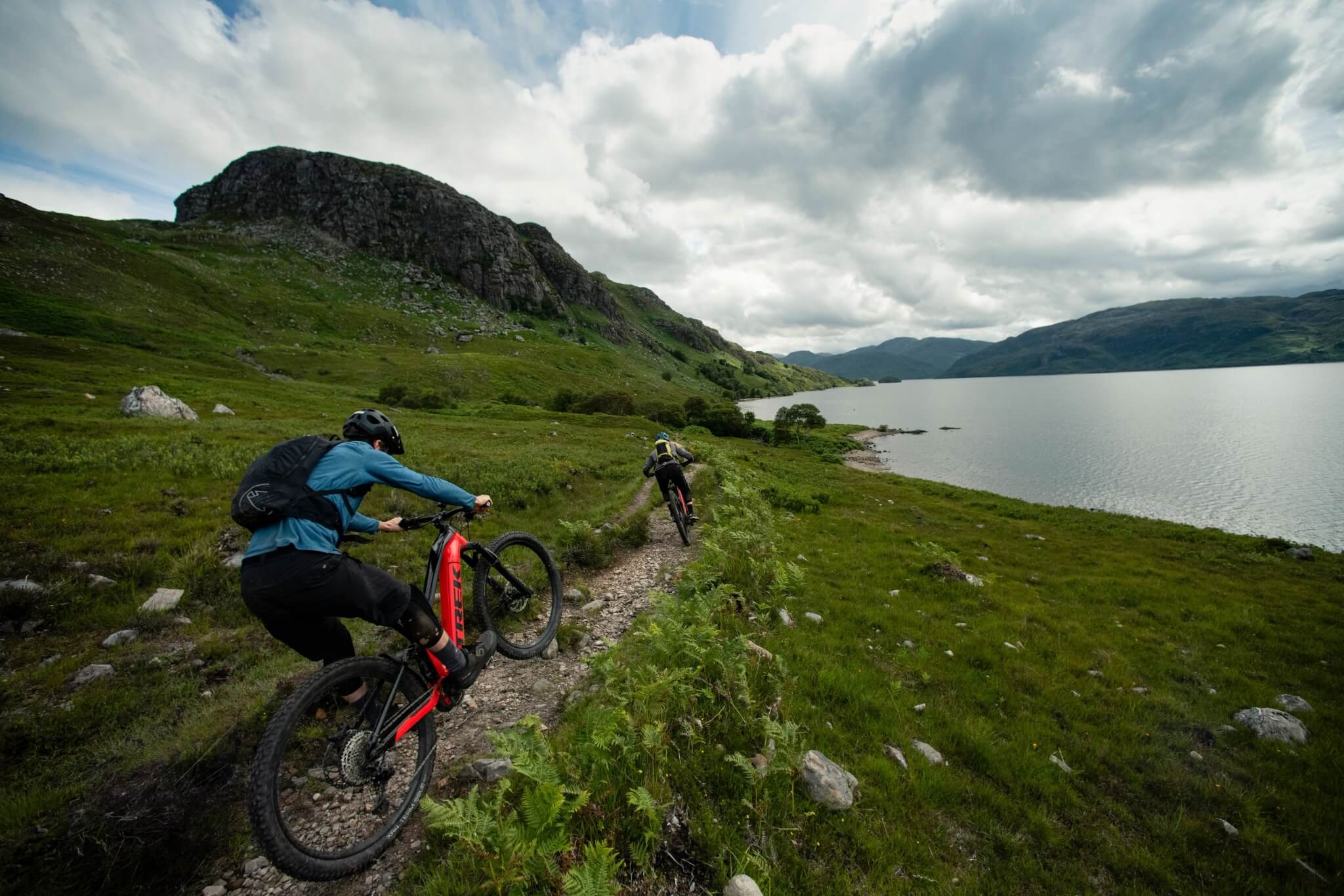Zwei Personen fahren Trek E-Mountainbikes auf einem Pfad entlang eines Sees in bergiger Landschaft.