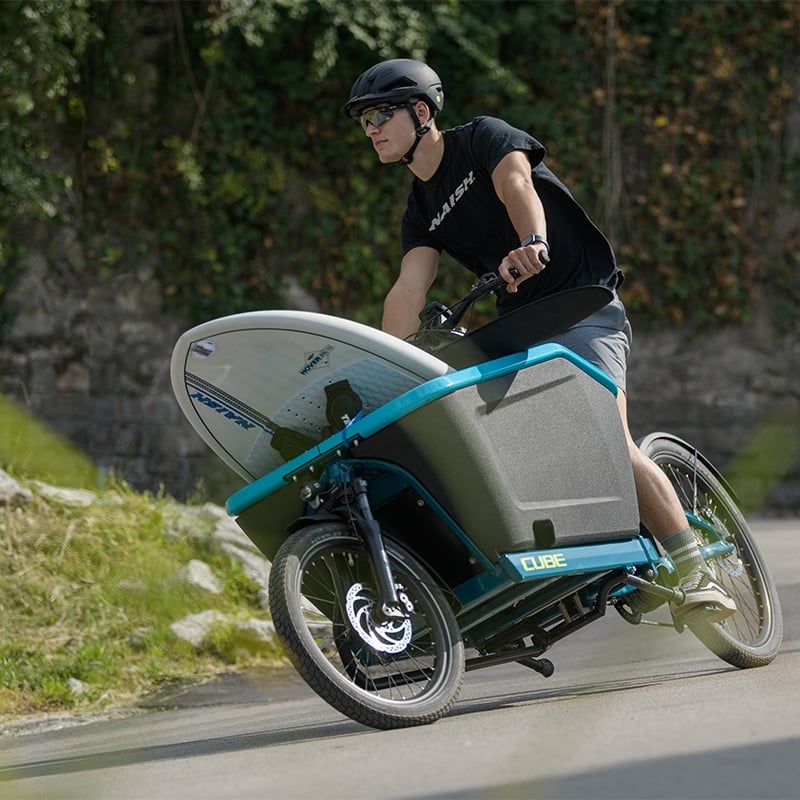 Mann auf einem Cube Cargo Hybrid Lastenrad transportiert ein Surfbrett, fährt auf einer Straße.