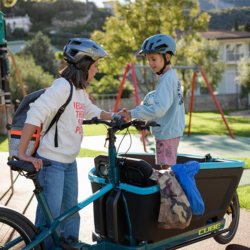 Eine Frau und ein Kind mit Helmen auf einem Cube Cargo Hybrid Lastenrad im Park.