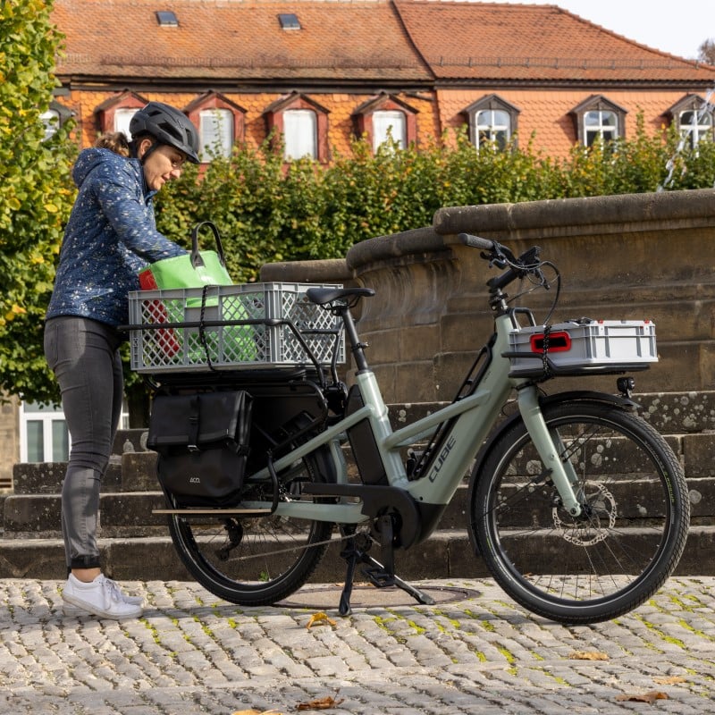 Person mit Helm lädt Einkäufe auf ein Cube Lastenrad vor einem Backsteingebäude. Ideal für Stadttransporte.