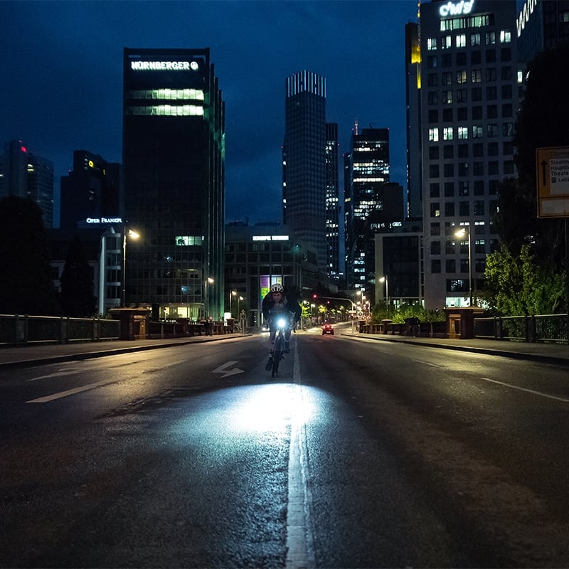Ein Radfahrer fährt bei Nacht durch eine beleuchtete Stadtstraße. Vorne eine starke Fahrradlampe.
